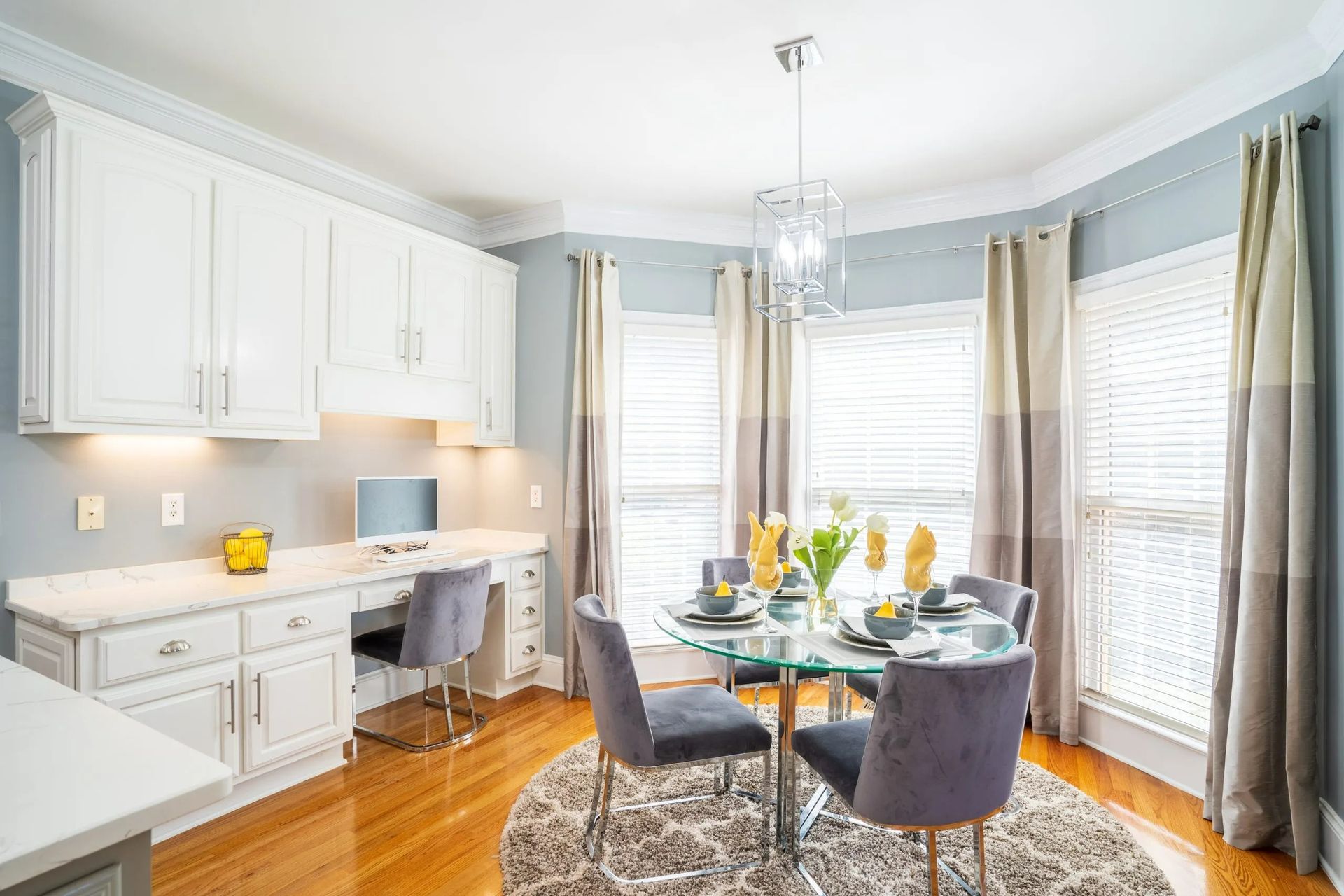 A bright home dining area with white cabinets, a desk workstation, and a glass-top table set for four with velvet chairs.