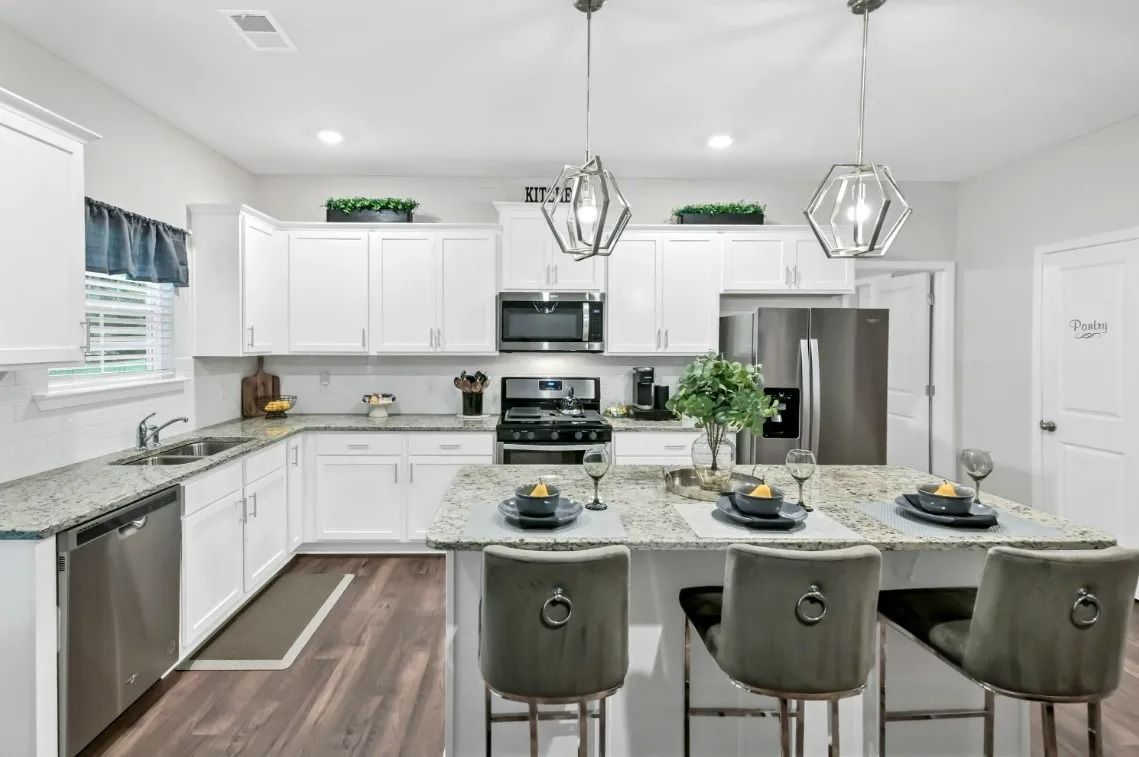 A modern white kitchen with an island, stone countertops, stainless steel appliances, and three gray bar stools.
