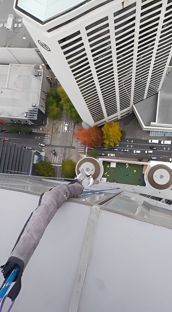 Person on building ledge, high angle view. Holding tools, with tall building and colorful trees below.