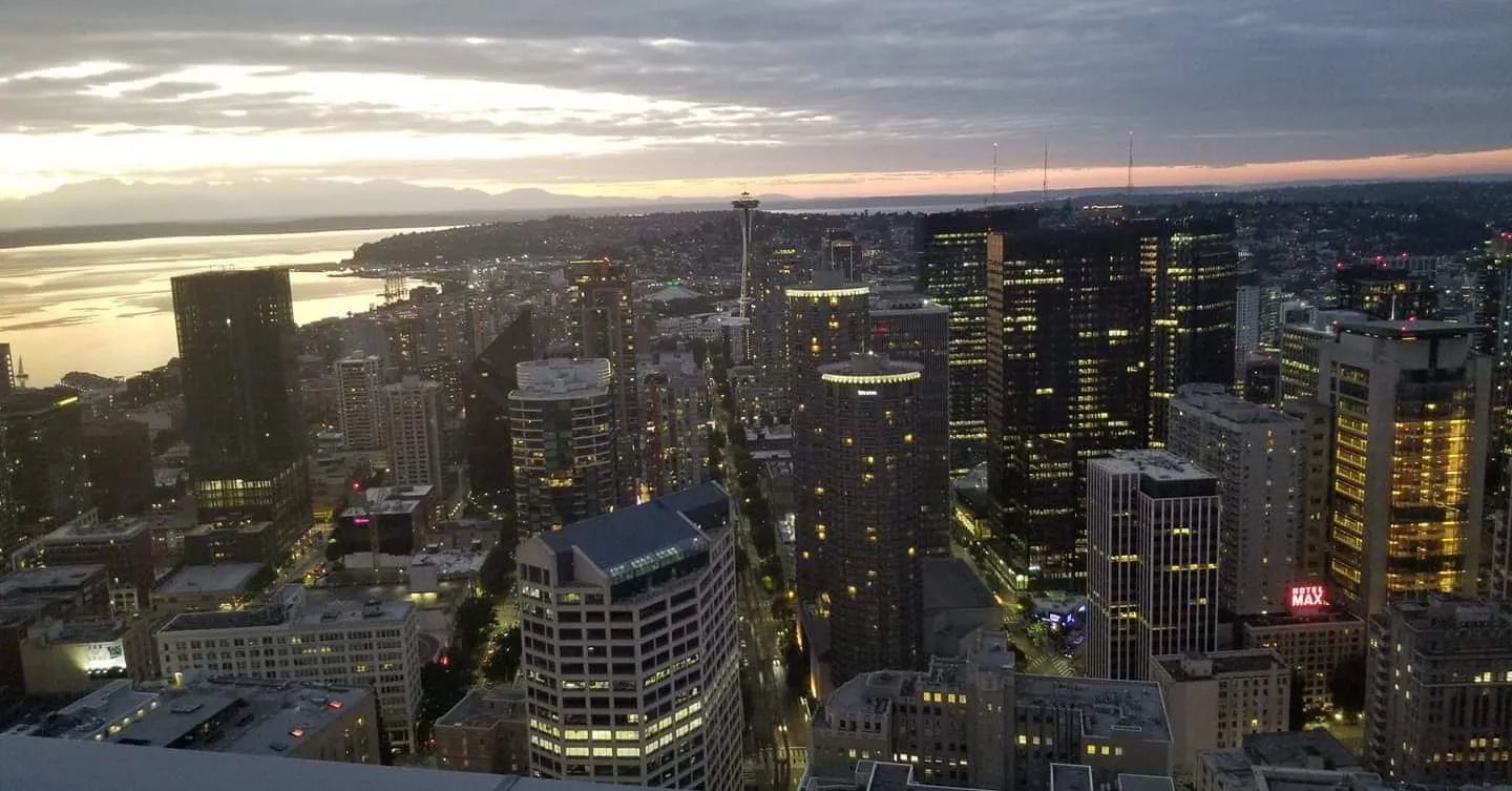 City skyline at dusk; lights twinkle. Buildings line a street towards a bay.