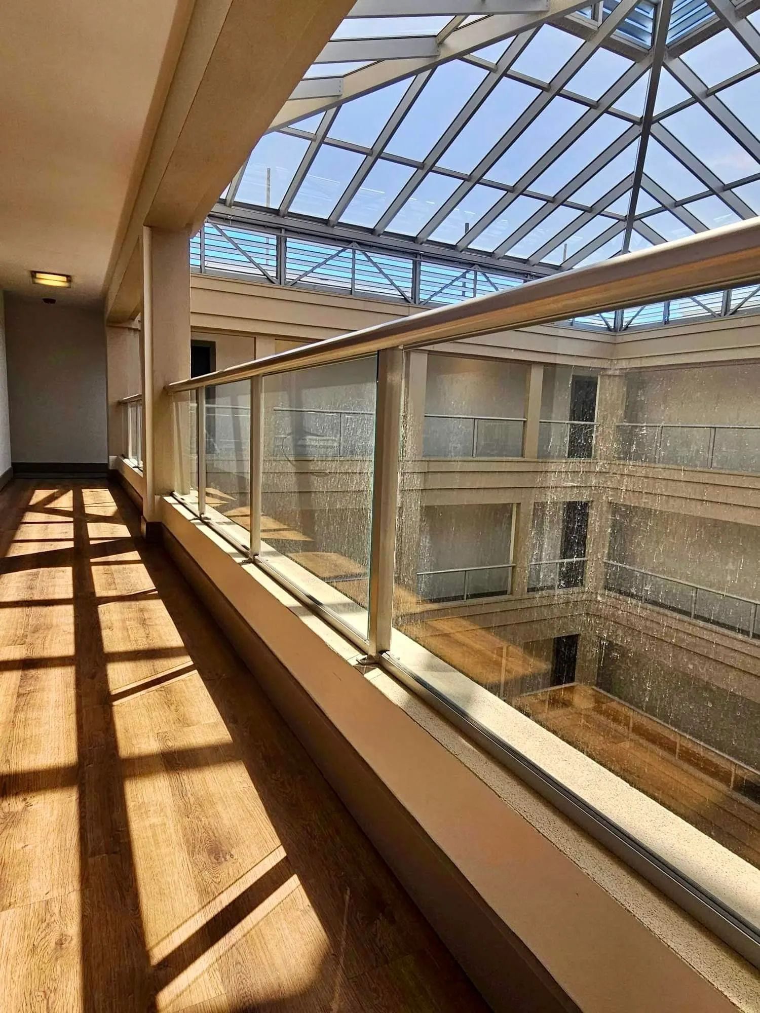 Hallway with glass railing, wooden floors, and skylight, with sunlight casting shadows.