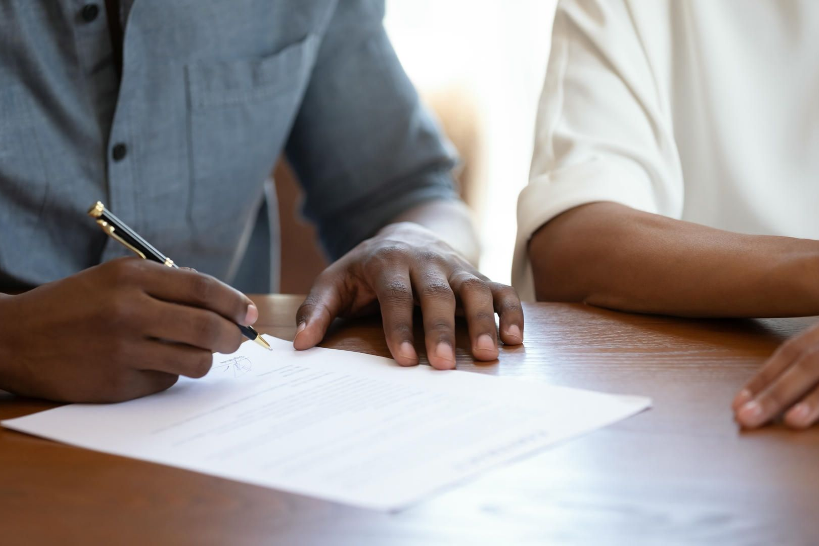 Person signing a document at a wooden table, next to another person with a white sleeve.