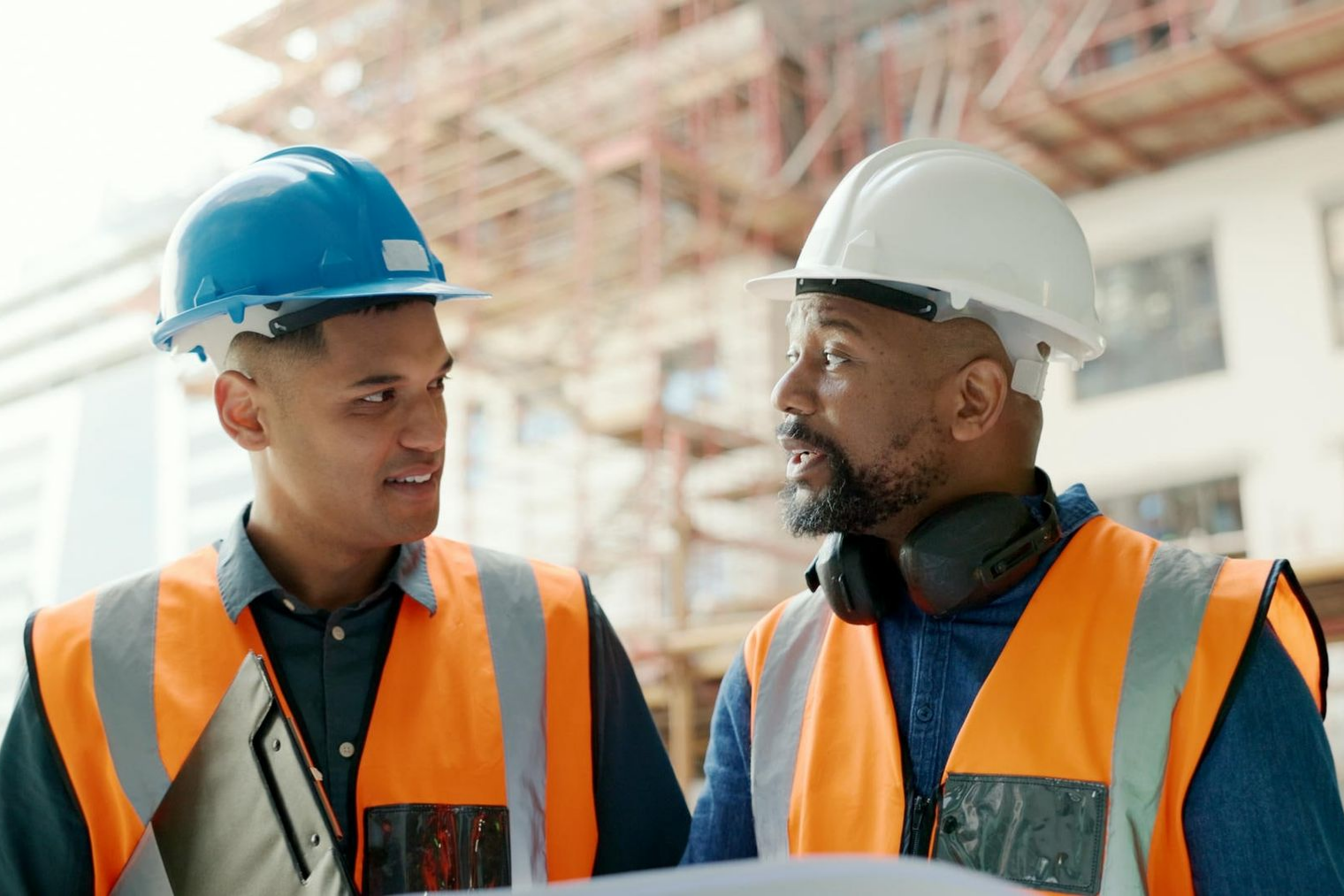 Two construction workers in hard hats and vests discussing blueprints on a construction site.