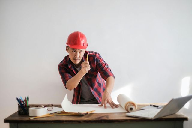 Una persona con casco rojo y camisa a cuadros señala unos planos sobre un escritorio