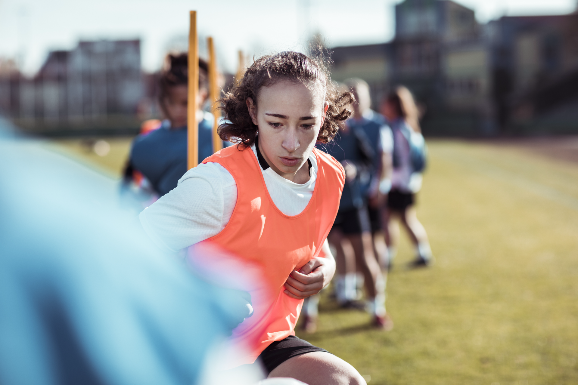 Girl in orange vest, running soccer drill on a green field. Others in blue nearby.