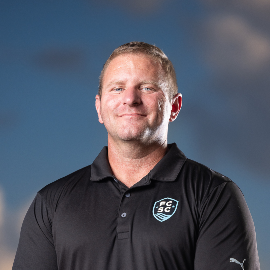 Man wearing a black polo shirt, smiling, with a logo on the left chest. Blue and white background.