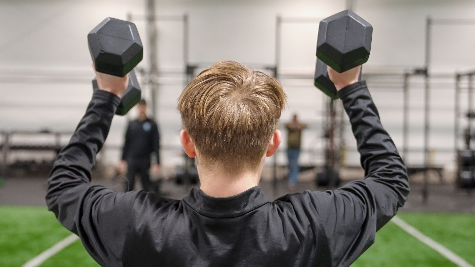 Person in black jacket lifting dumbbells overhead in a gym.