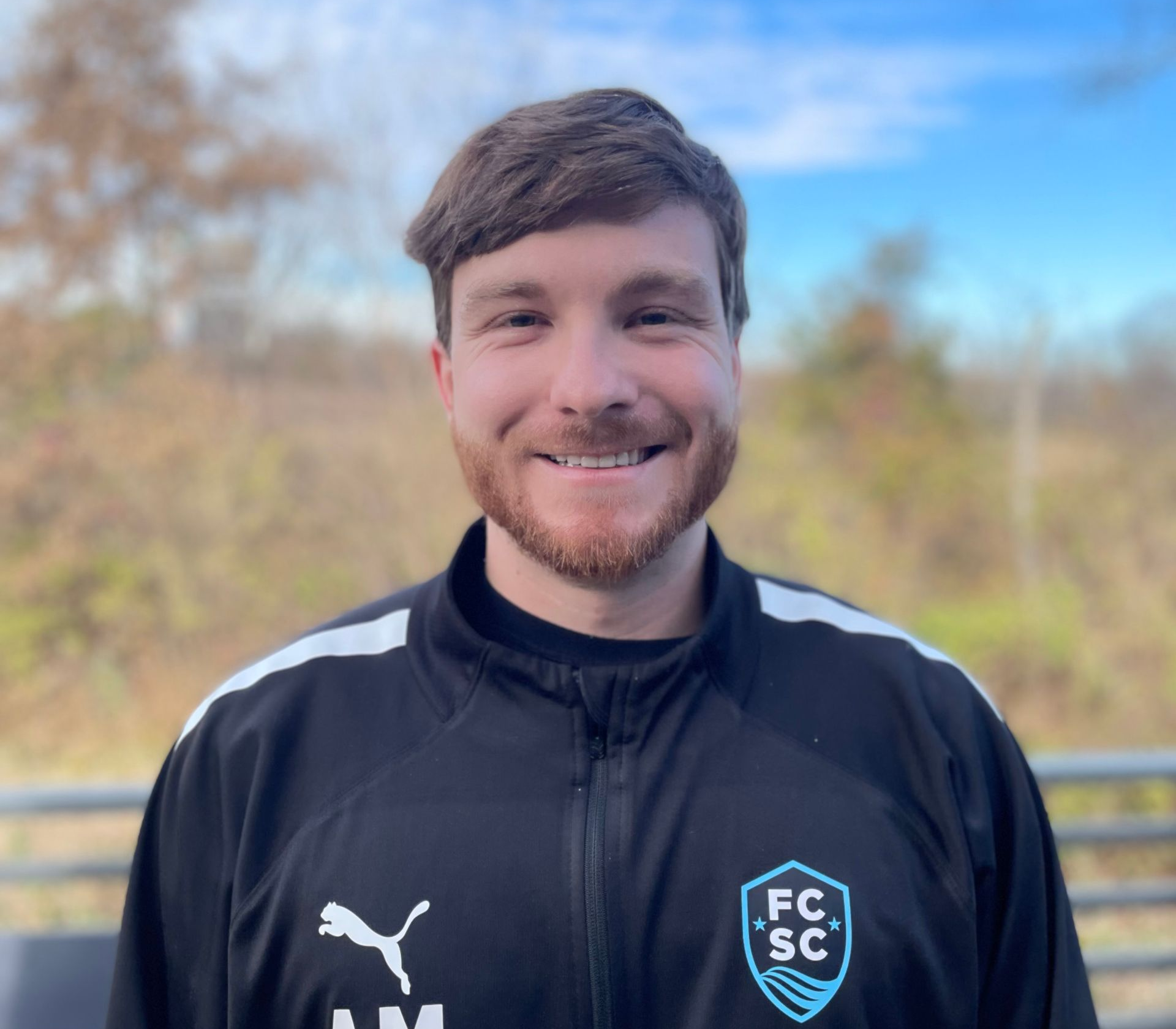 Man wearing a black polo shirt, smiling, with a logo on the left chest. Blue and white background.