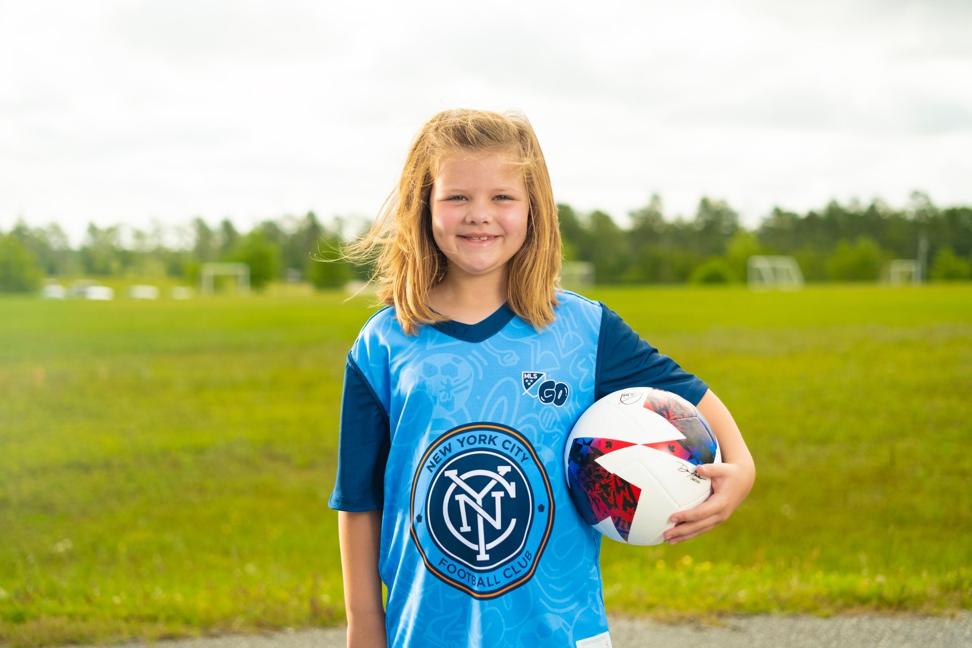 Girl in black soccer uniform runs toward a soccer ball on a green field. Indoor setting.