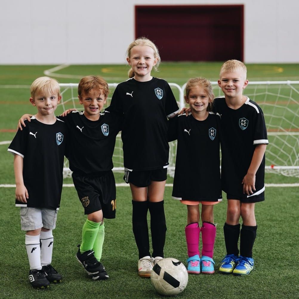 Five children in black soccer uniforms stand on a green field, arm in arm, with a soccer ball.