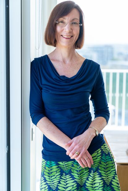 Woman with short brown hair, wearing glasses, blue top and green patterned skirt, leaning against a window, smiling.