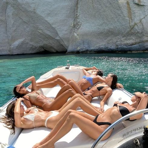 Six women in bikinis sunbathe on a boat in clear, turquoise water. White cliffs are in the background.