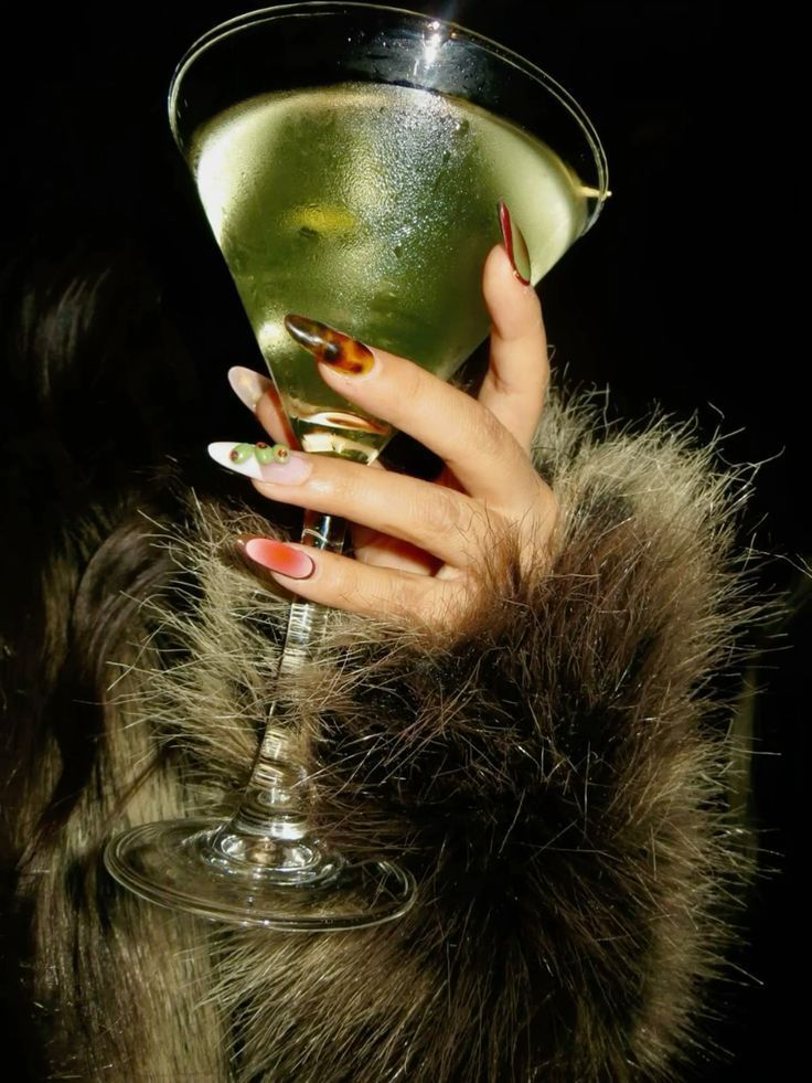Hand with long, decorated nails holding a cocktail glass, against a fluffy, brown fur sleeve.
