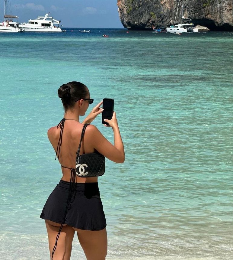 Woman on a beach taking a photo with a phone. Wearing a black outfit, sunglasses, and a purse, ocean and boats in the background.