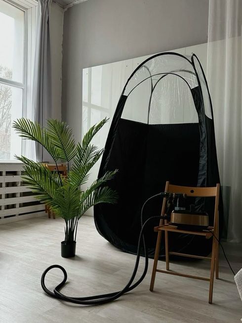 A tanning tent setup in a room with a plant and equipment on a chair.
