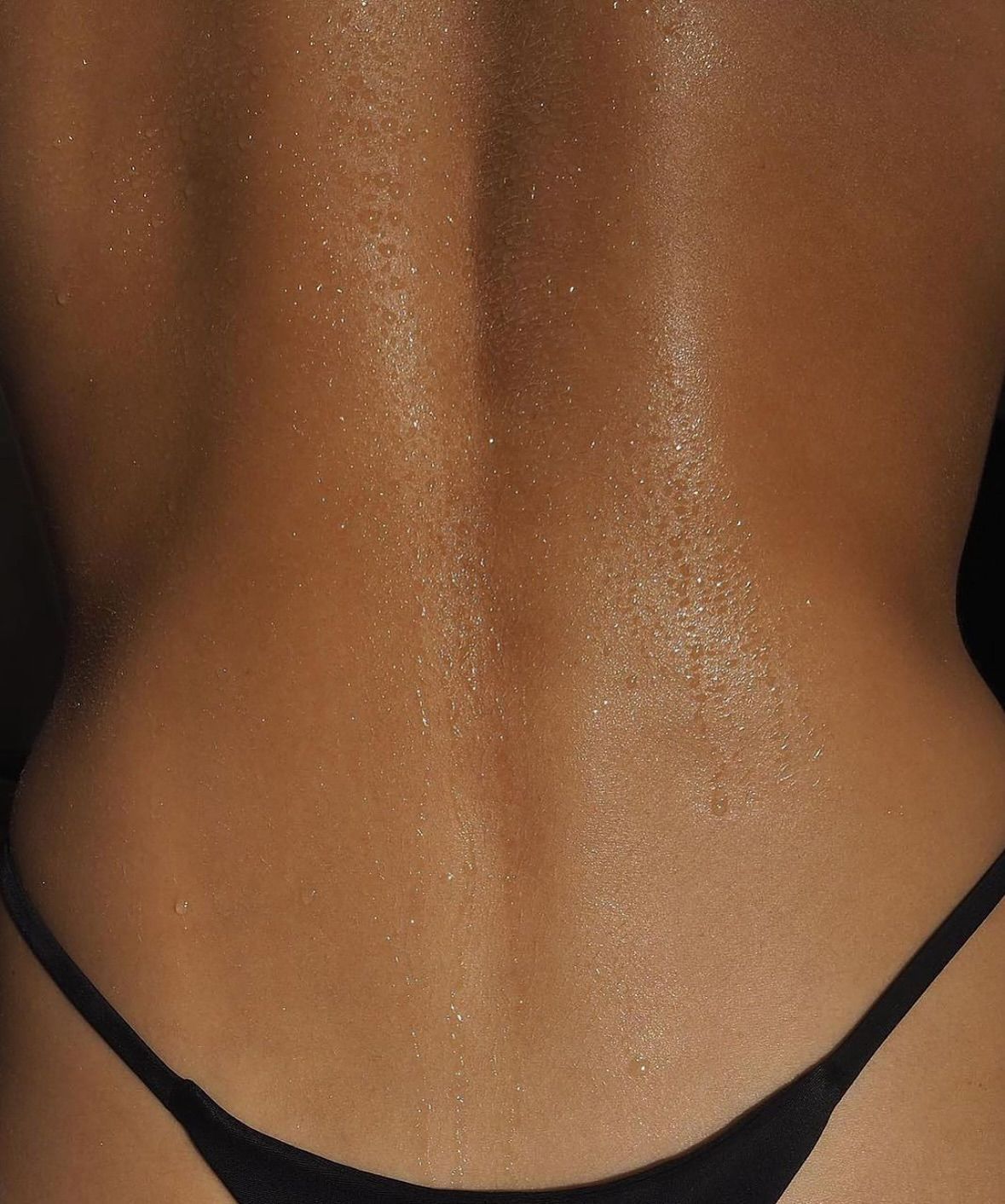 Back of a person glistening with oil. Dark skin, black bikini bottom.