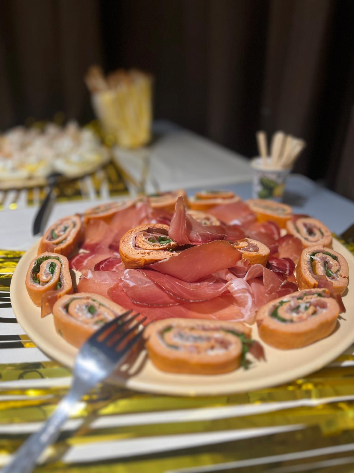 Piatto di involtini di salmone, prosciutto e antipasti su un tavolo, con una forchetta in primo piano.