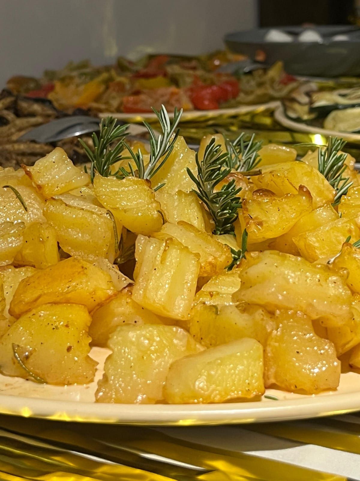 Patate arrosto guarnite con rosmarino su un piatto da portata, con un piatto di verdure sullo sfondo.