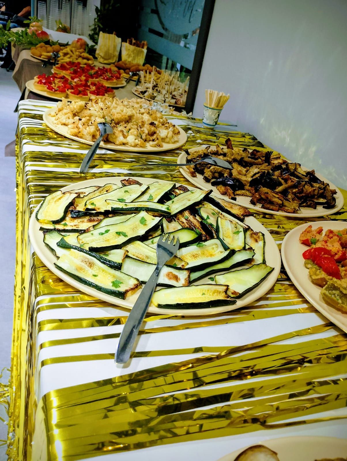 Un tavolo di cibo preparato al momento: zucchine, melanzane, pasta, bruschette su una tovaglia con frange dorate.