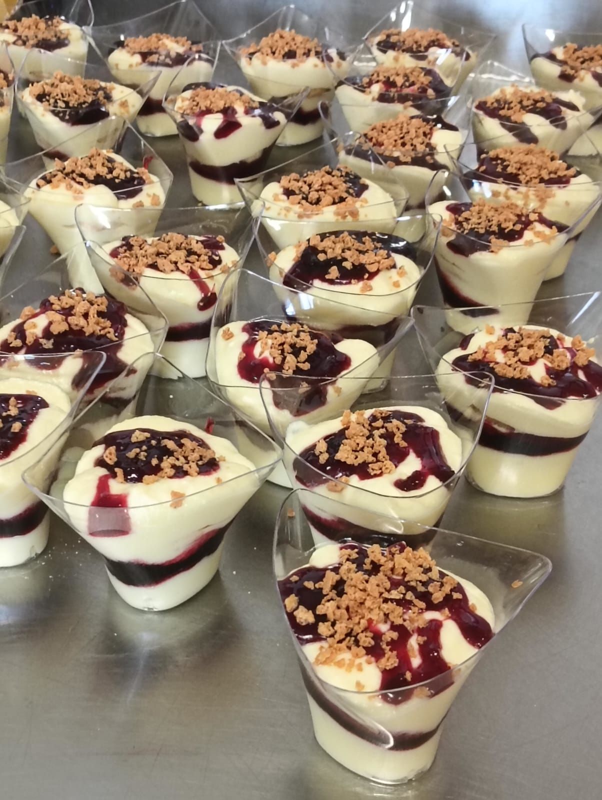 Piccoli dessert in tazze trasparenti, ricoperti di crema bianca, salsa ai frutti di bosco e briciole.