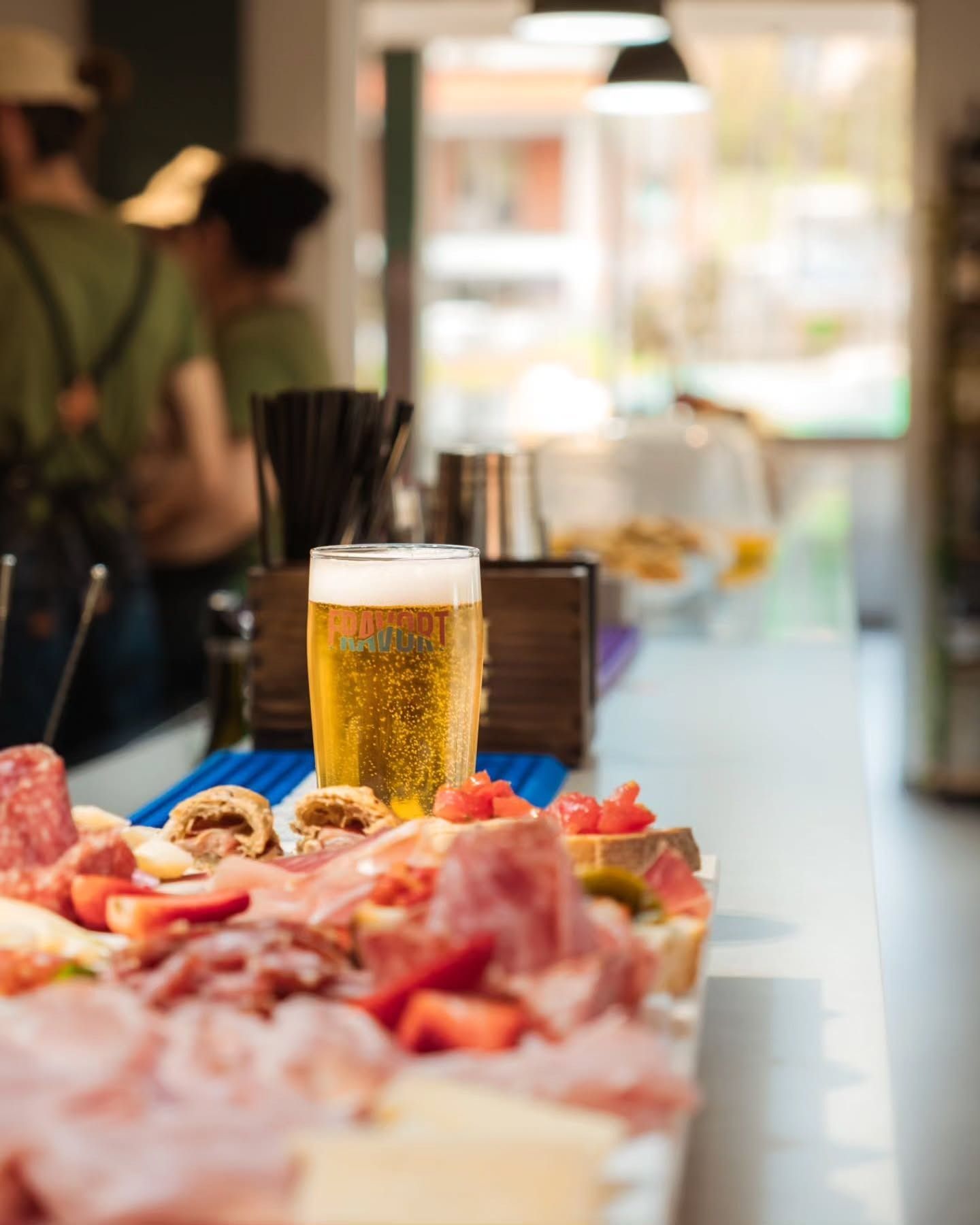 Un bicchiere di birra e un tagliere di salumi su un bancone del bar. Persone e una finestra sullo sfondo.