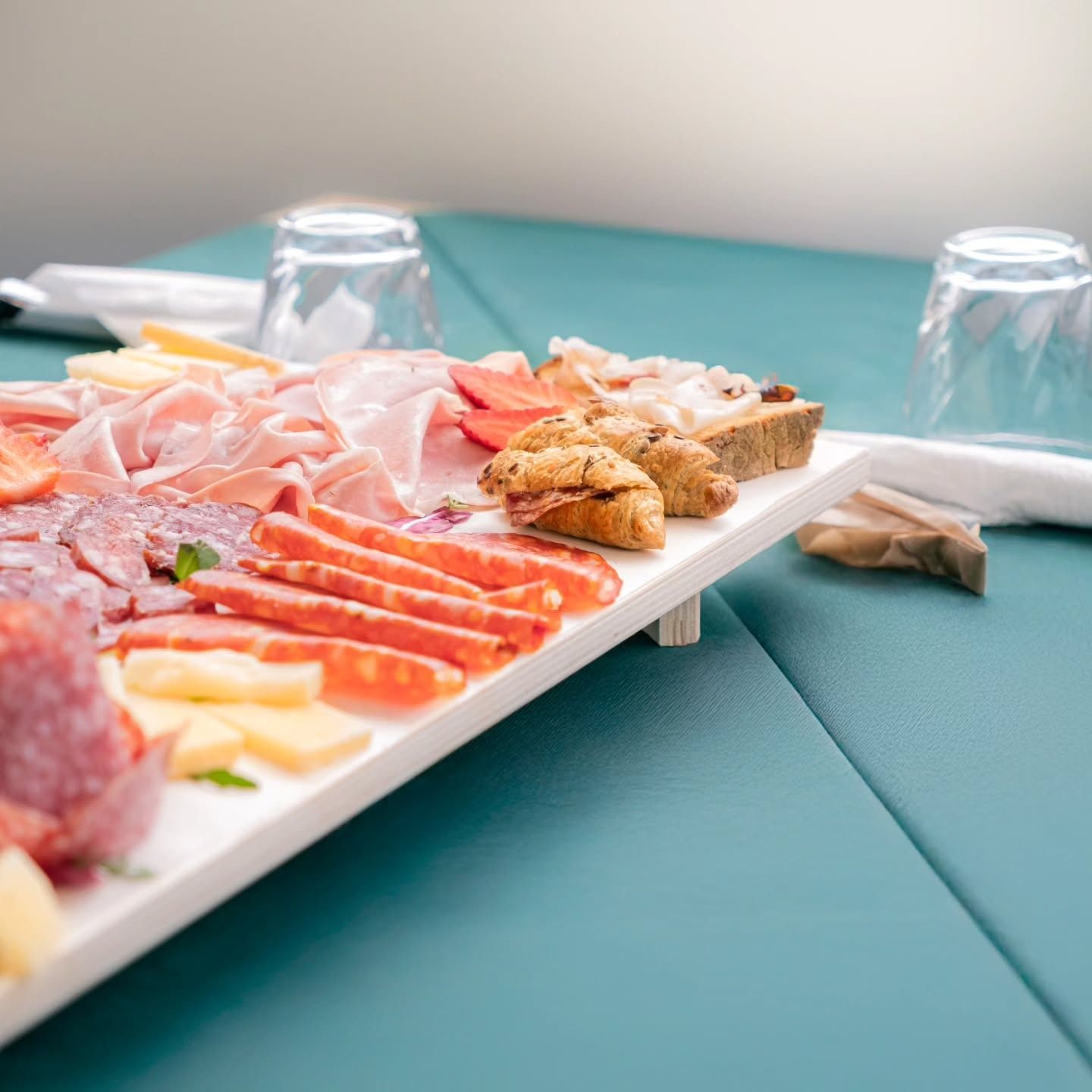 Tagliere di salumi, formaggi e pane su un tavolo verde acqua con bicchieri.
