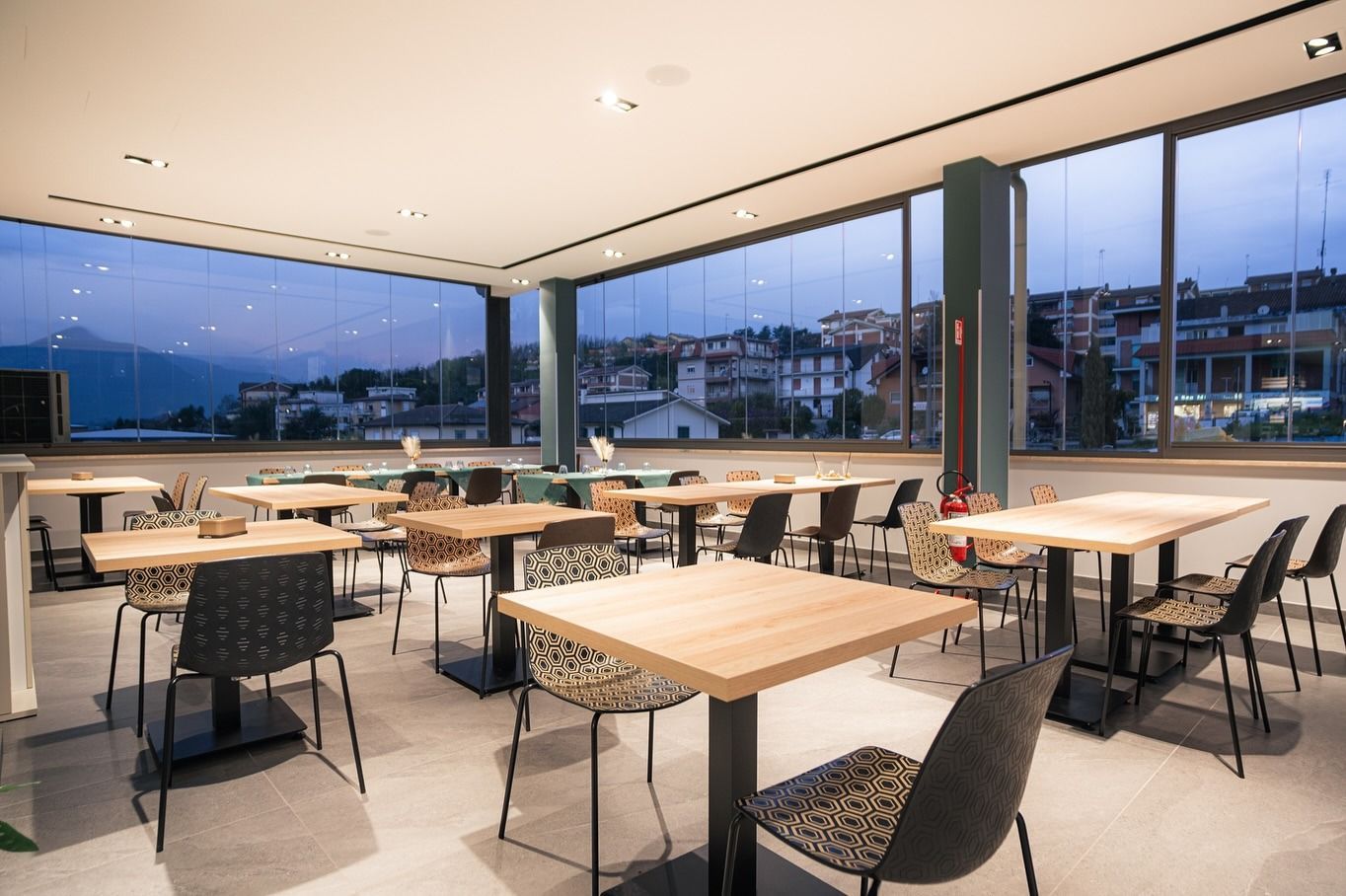 Interno vuoto di un ristorante con tavoli quadrati, sedie e grandi finestre con vista sulla città e sull'acqua.