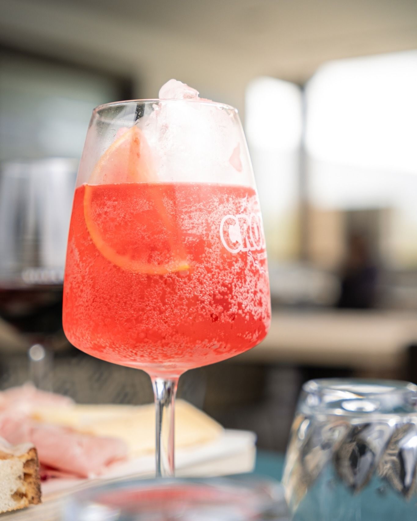 Cocktail rosso in un bicchiere alto con ghiaccio e fetta d'arancia, vicino al cibo e a un bicchiere d'acqua.
