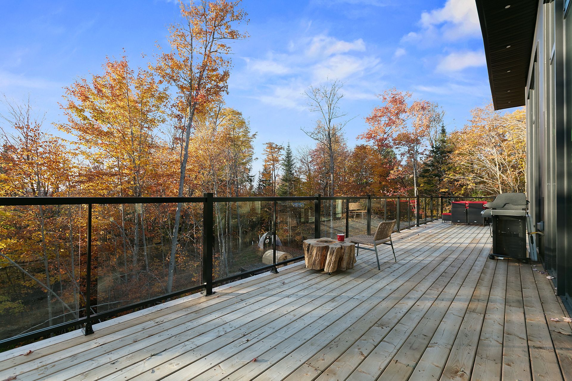 Terrasse en bois donnant sur des arbres d'automne, rambarde en verre, petite table avec du bois de chauffage.