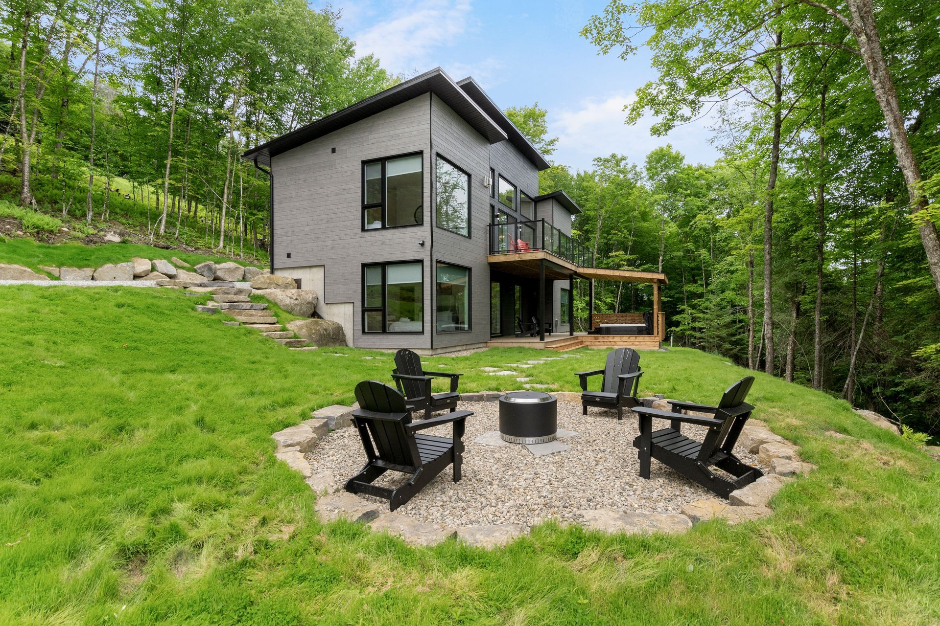 Maison grise moderne avec des chaises noires autour d'un foyer sur une colline herbeuse, entourée d'arbres.