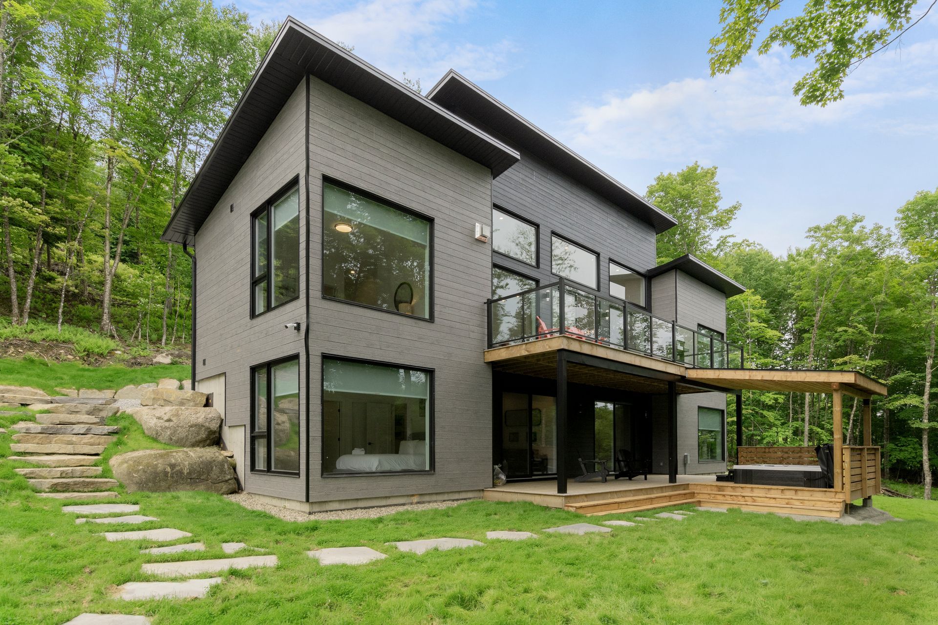 Maison grise moderne avec de grandes fenêtres, un balcon et un chemin de pierre dans un espace herbeux arboré.