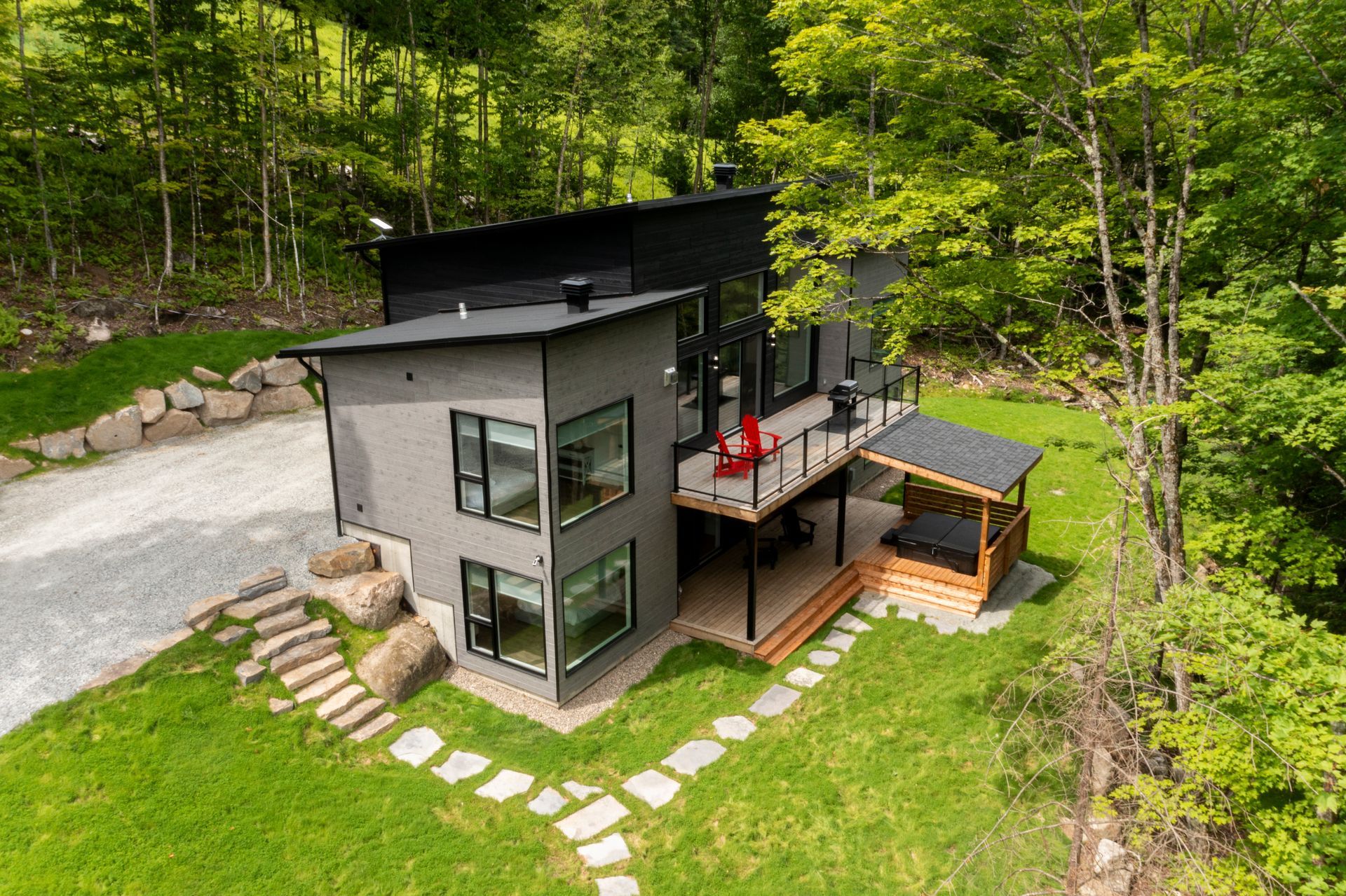 Maison moderne à deux étages avec bardage gris foncé, toit noir et terrasse en bois entourée d'arbres.