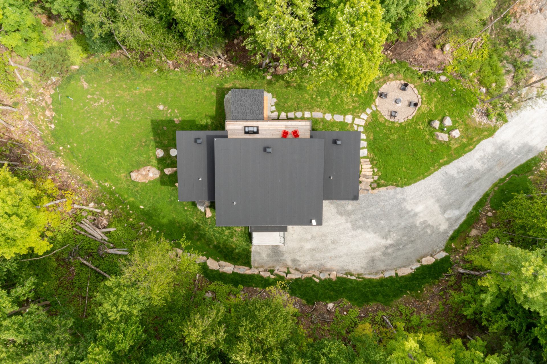 Vue aérienne d'une maison moderne avec un toit sombre et une allée de gravier, entourée d'arbres et de pelouse.