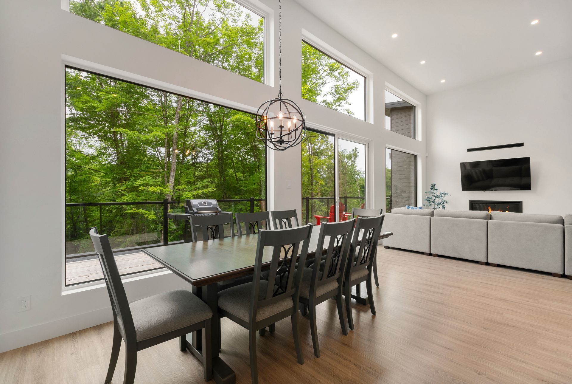 Salle à manger avec de grandes fenêtres, table et chaises, et un lustre, offrant une vue sur les arbres.