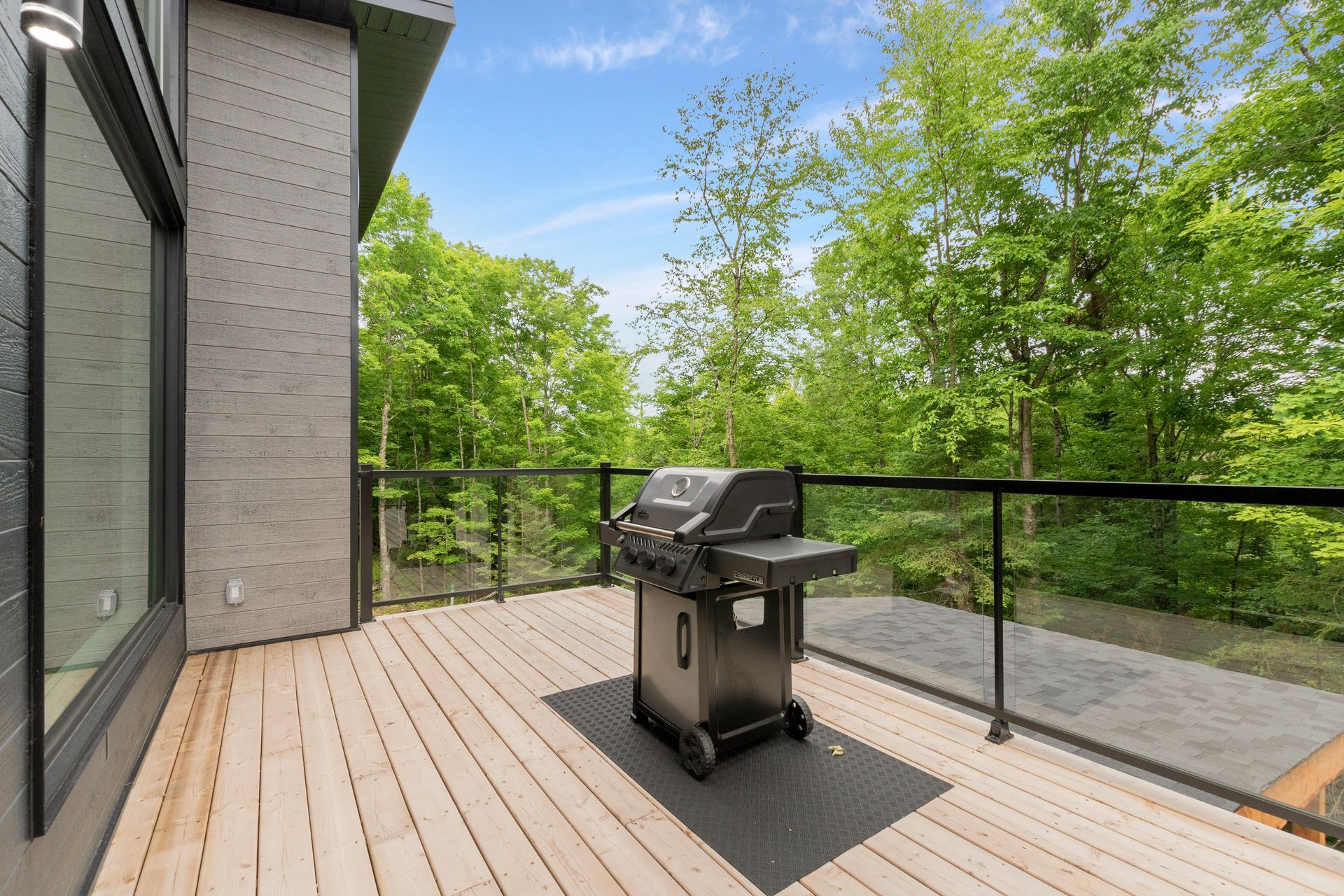 Terrasse avec barbecue noir, rambarde en verre et vue sur la forêt par une journée ensoleillée.
