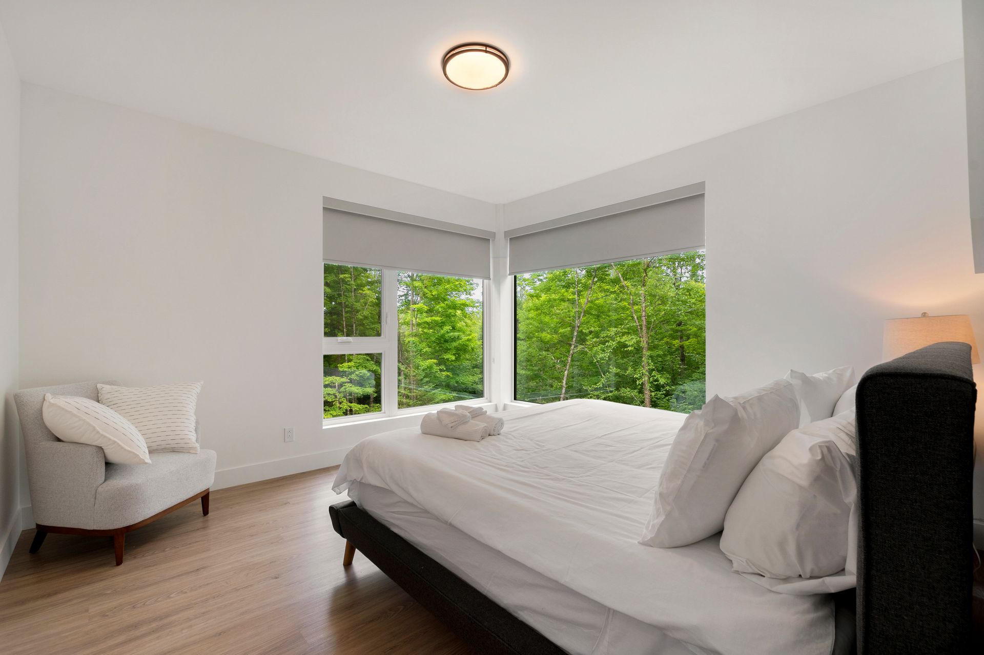 Chambre avec un lit, une petite chaise et des fenêtres donnant sur la verdure ; murs clairs et parquet.