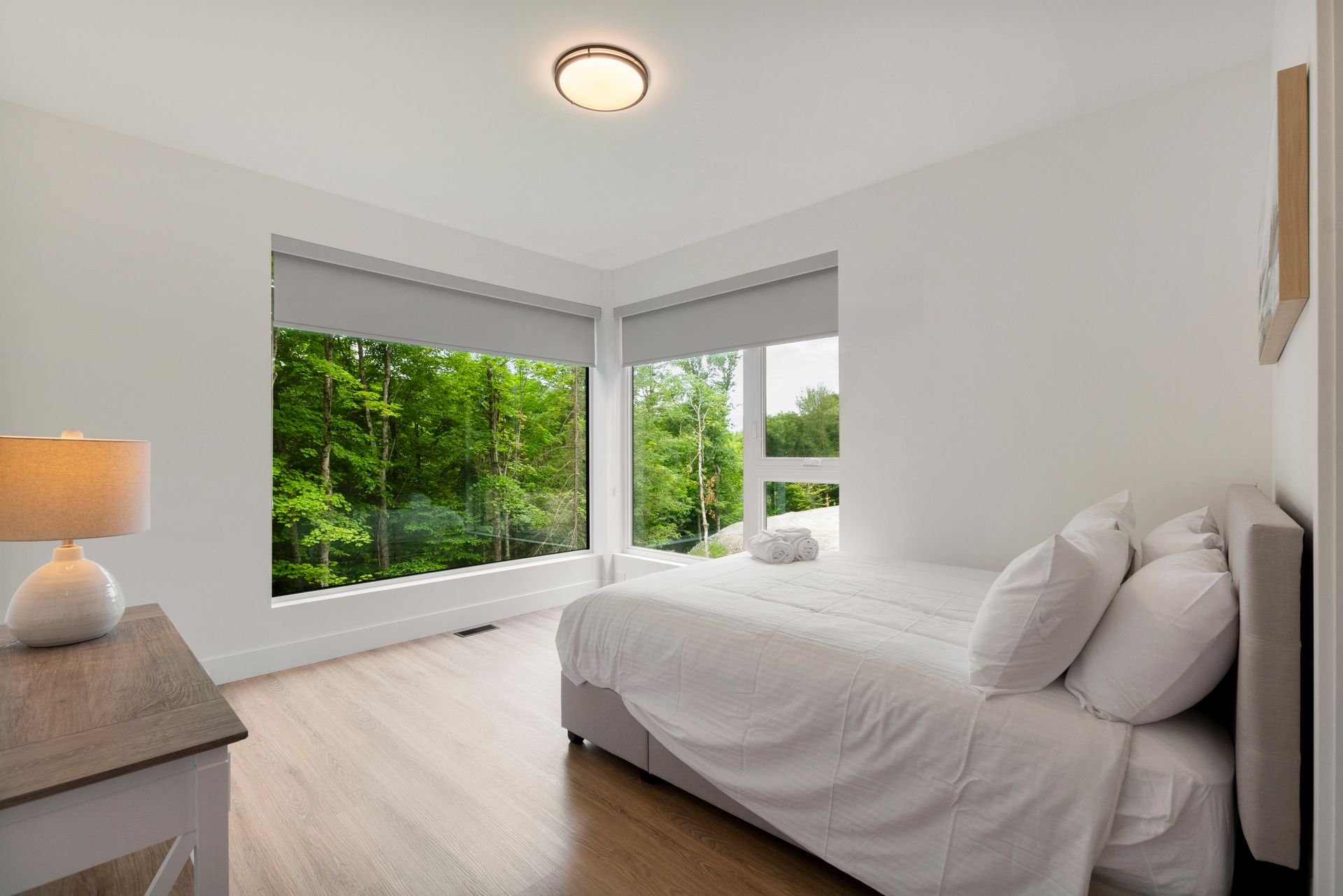 Chambre avec de grandes fenêtres donnant sur une forêt verdoyante luxuriante, literie blanche, table de chevet et plafonnier.