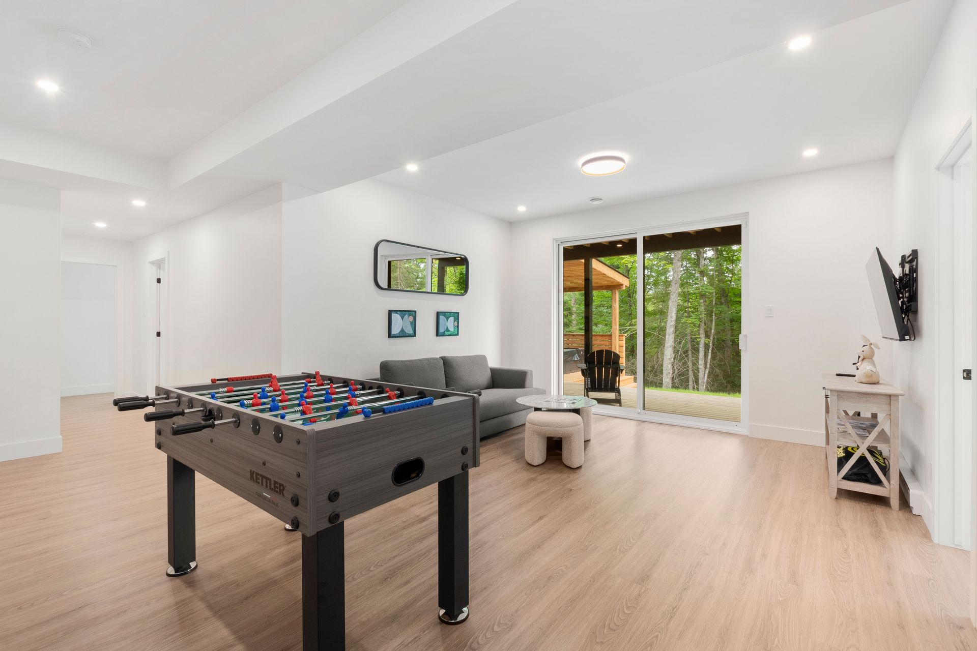 Sous-sol moderne avec baby-foot, canapé et portes-fenêtres coulissantes donnant sur une terrasse. Tons clairs et neutres.