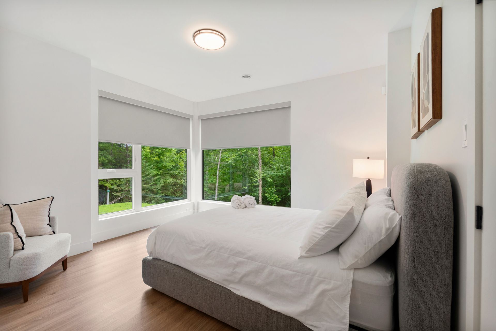 Chambre lumineuse avec un lit, une petite chaise et de grandes fenêtres donnant sur la verdure.