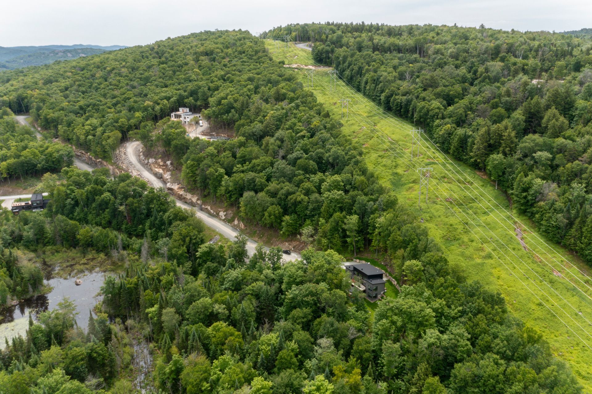 Colline verdoyante traversée par un large chemin dégagé ; une route serpente à proximité. La forêt entoure le chemin, une petite construction est visible.