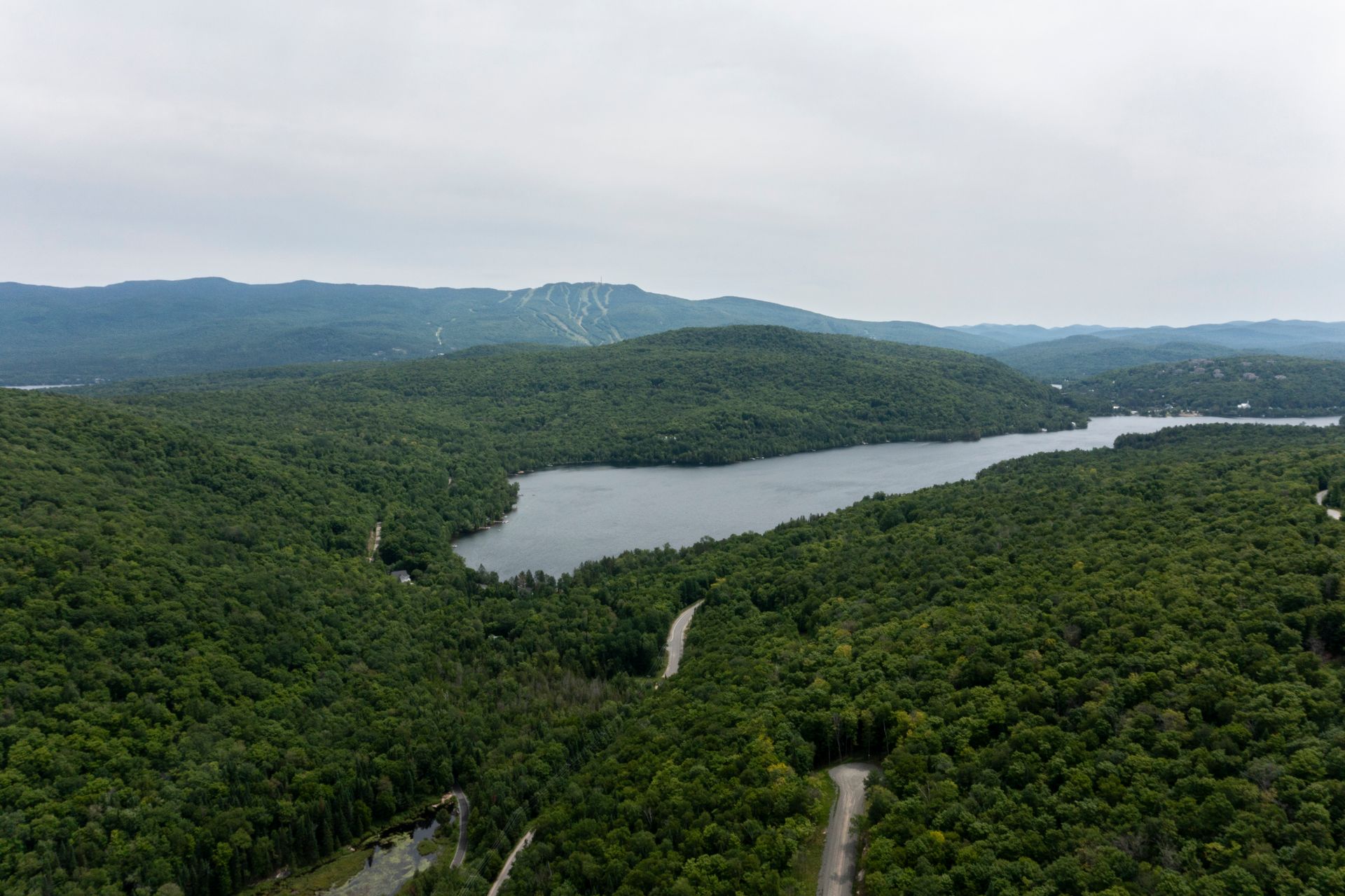 Vue aérienne d'un lac entouré de forêts verdoyantes et de collines ondulantes sous un ciel nuageux.