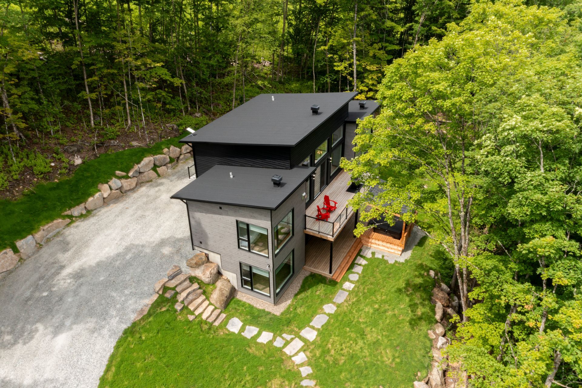 Maison moderne à deux étages avec toit et bardage gris foncé, nichée dans un quartier boisé. Allée en gravier et chemin de pierre.