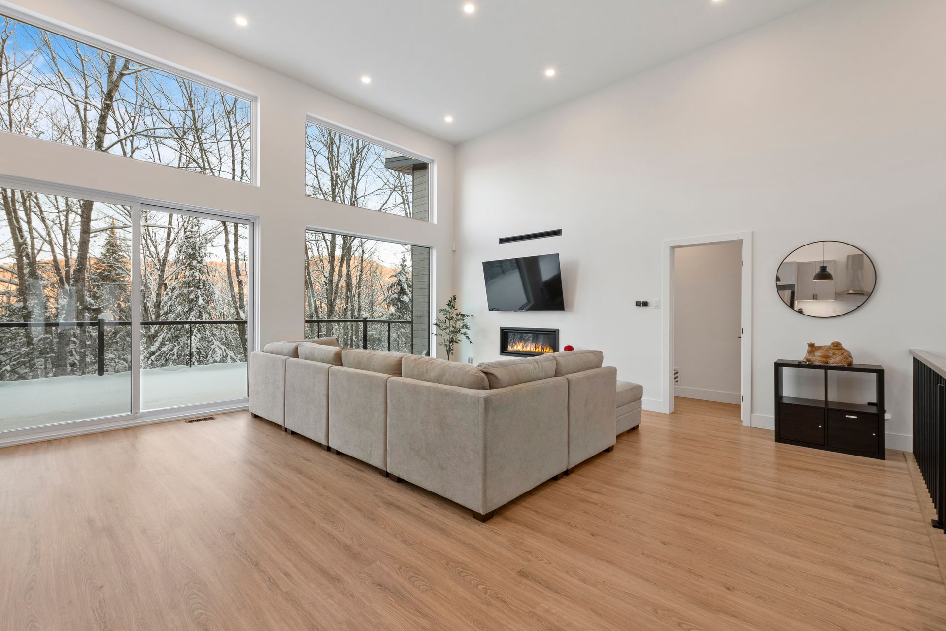 Salon moderne avec de grandes fenêtres donnant sur une forêt enneigée. Canapé clair, cheminée et parquet.