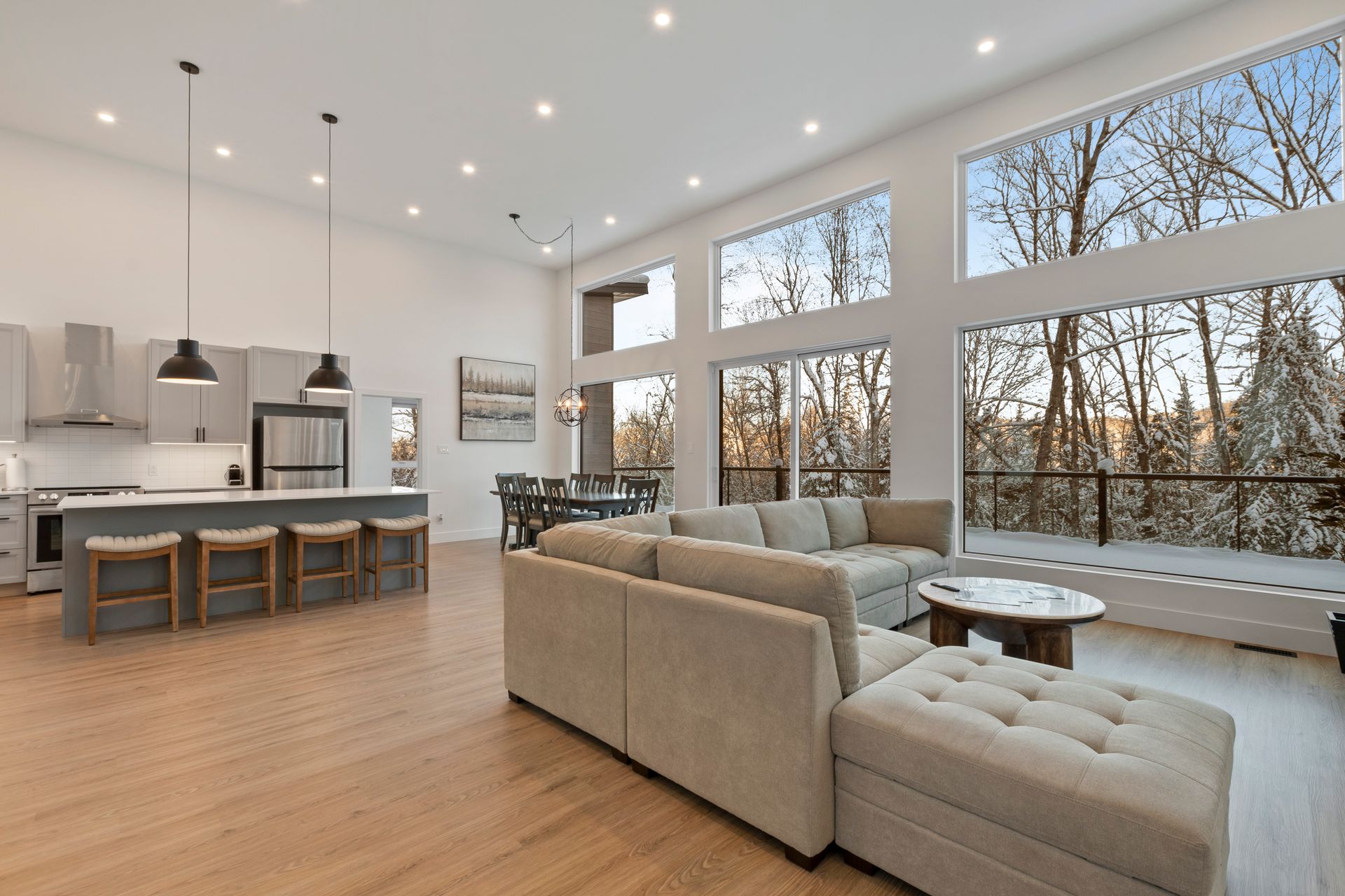Espace de vie à aire ouverte comprenant une cuisine, un coin repas et de grandes fenêtres donnant sur une vue boisée enneigée.