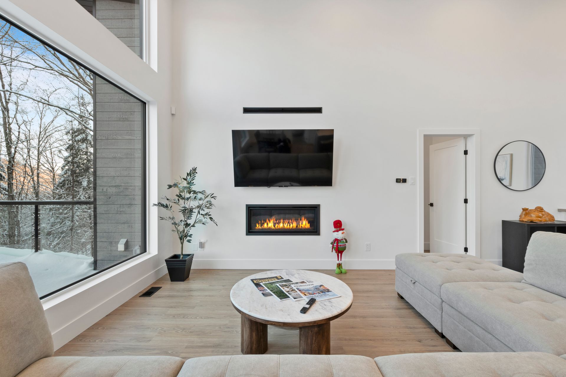 Salon avec cheminée, télévision, grande fenêtre, canapé et table basse ronde décorée ; tons neutres.