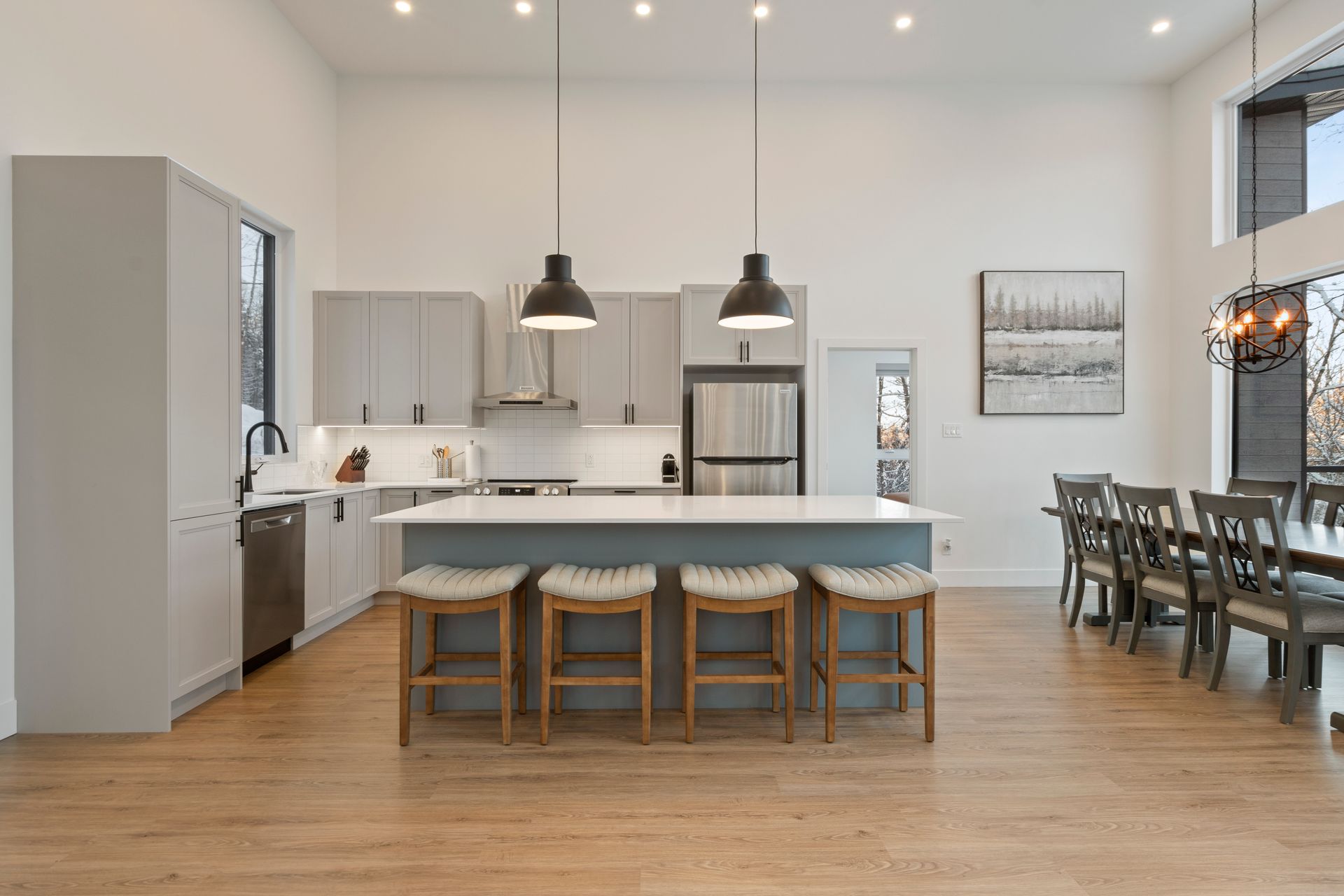 Cuisine moderne avec îlot central, coin repas et coin salon ; couleurs neutres, hauts plafonds, suspensions lumineuses.