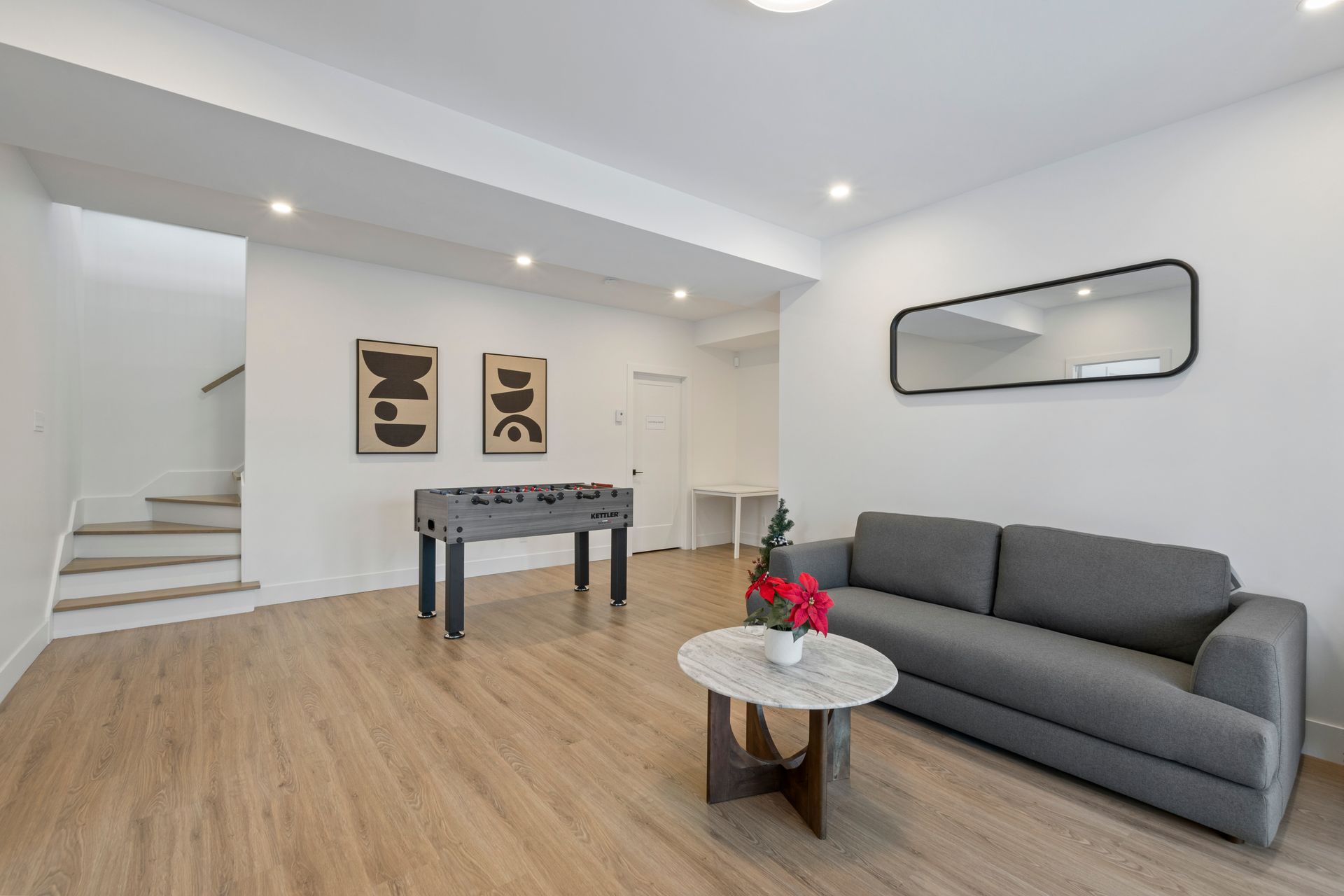 Espace de vie moderne au sous-sol, canapé gris, baby-foot, œuvres d'art, escalier et miroir.