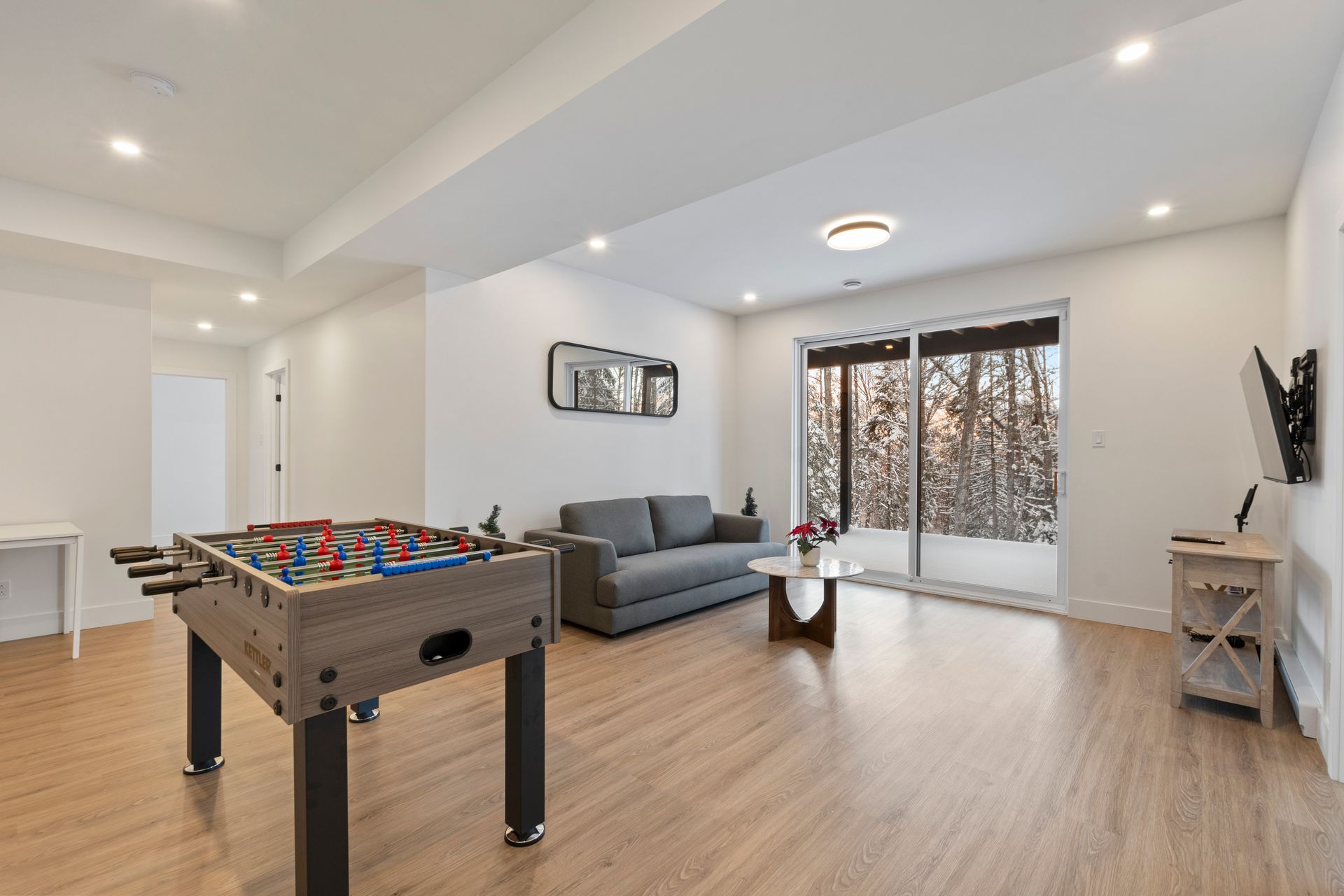 Table de baby-foot dans un salon moderne avec canapé, télévision et porte-fenêtre coulissante.