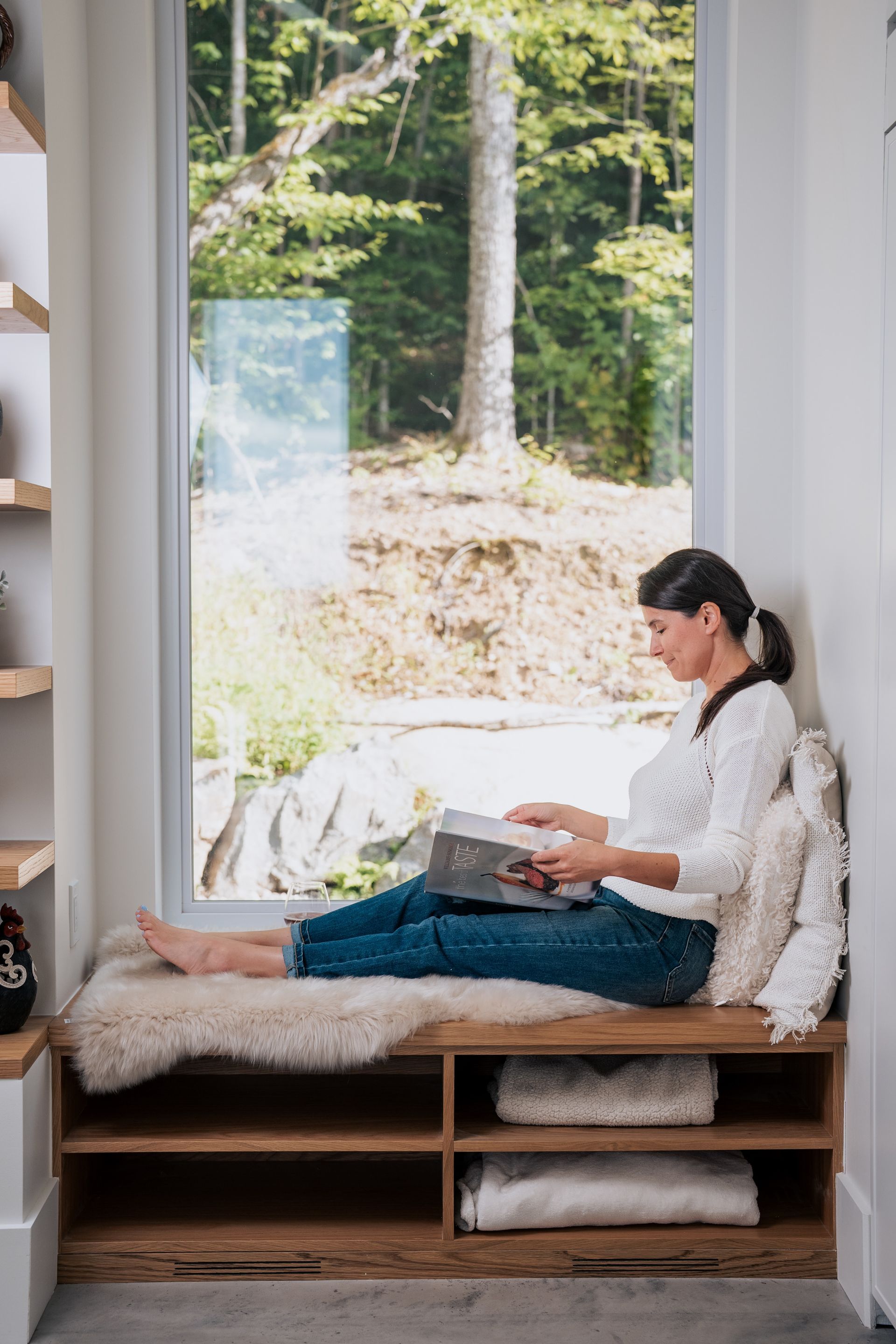 Femme lisant sur une banquette près d'une fenêtre avec vue sur les arbres, vêtue d'un pull blanc et d'un jean.