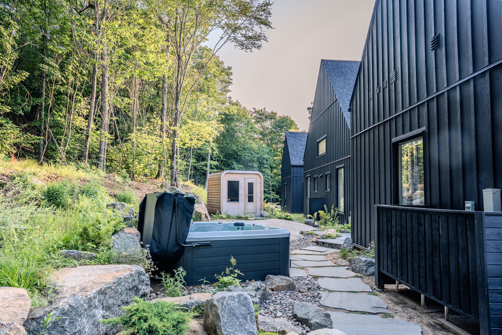 Cabanes noires avec chemin de pierre, bain à remous et petite dépendance dans une zone boisée.